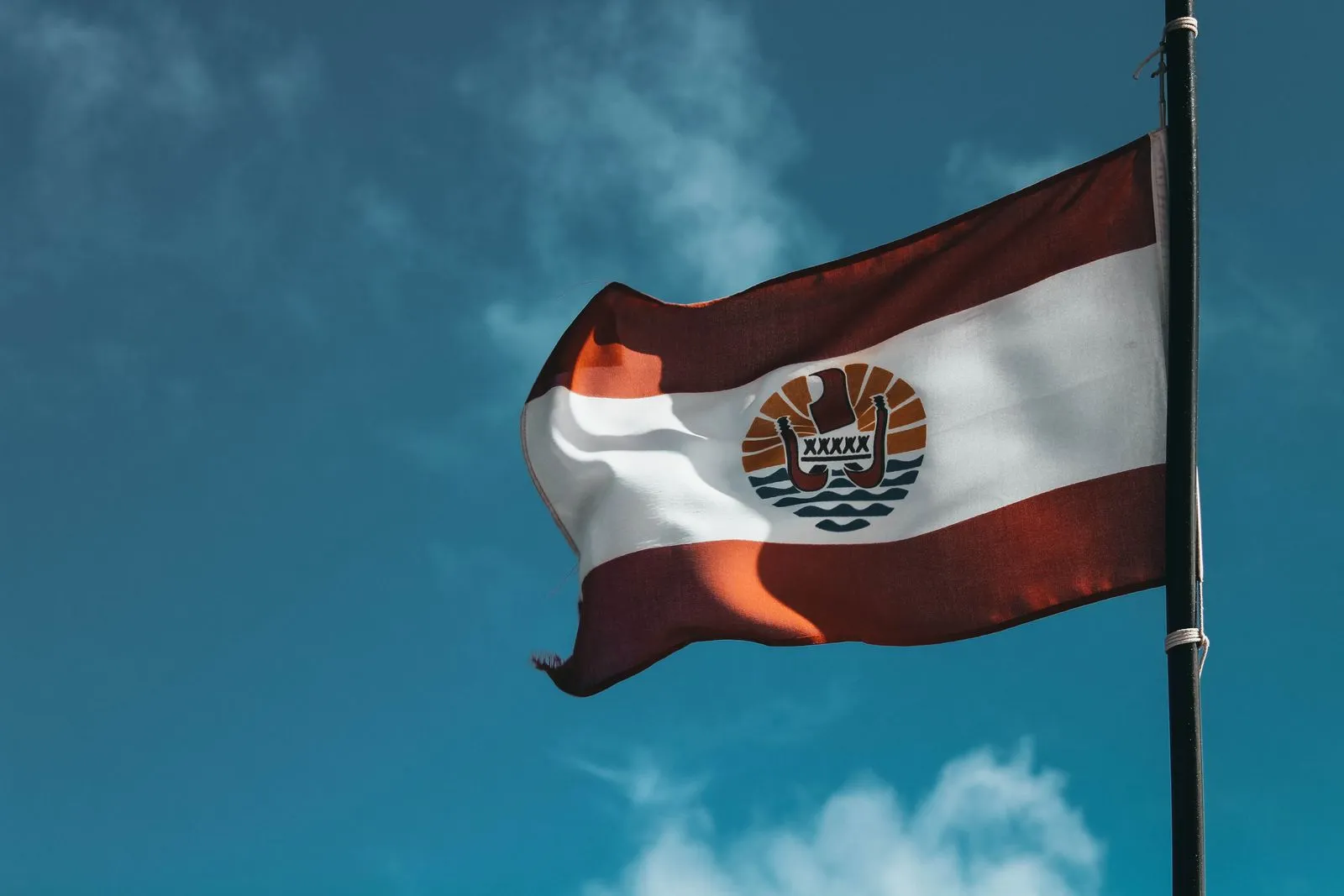 Drapeau de la Polynésie française hissé sur son mât, bandes rouge et blanche et emblème à la pirogue