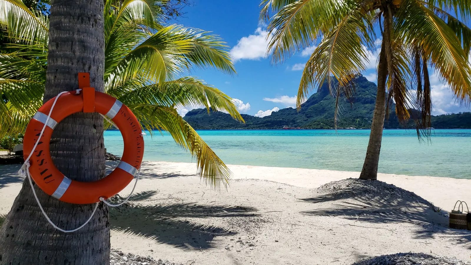 Plage de sable blanc ombragée de cocotiers, probablement sur un motu, avec le lagon turquoise et le relief de Bora Bora à l'horizon