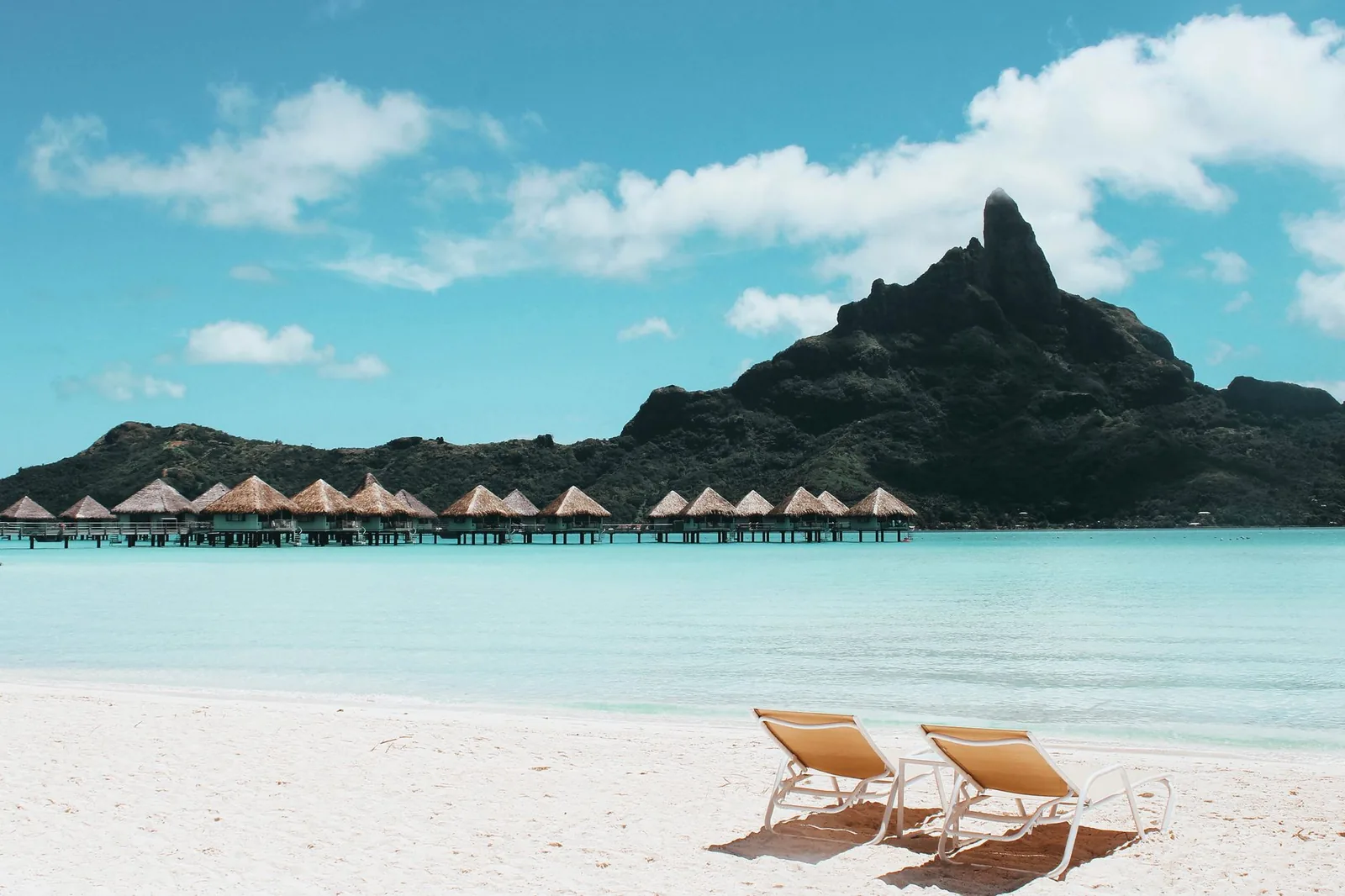 Plage de sable blanc et transats face au lagon turquoise de Bora Bora, avec le mont Otemanu et des bungalows sur pilotis au fond