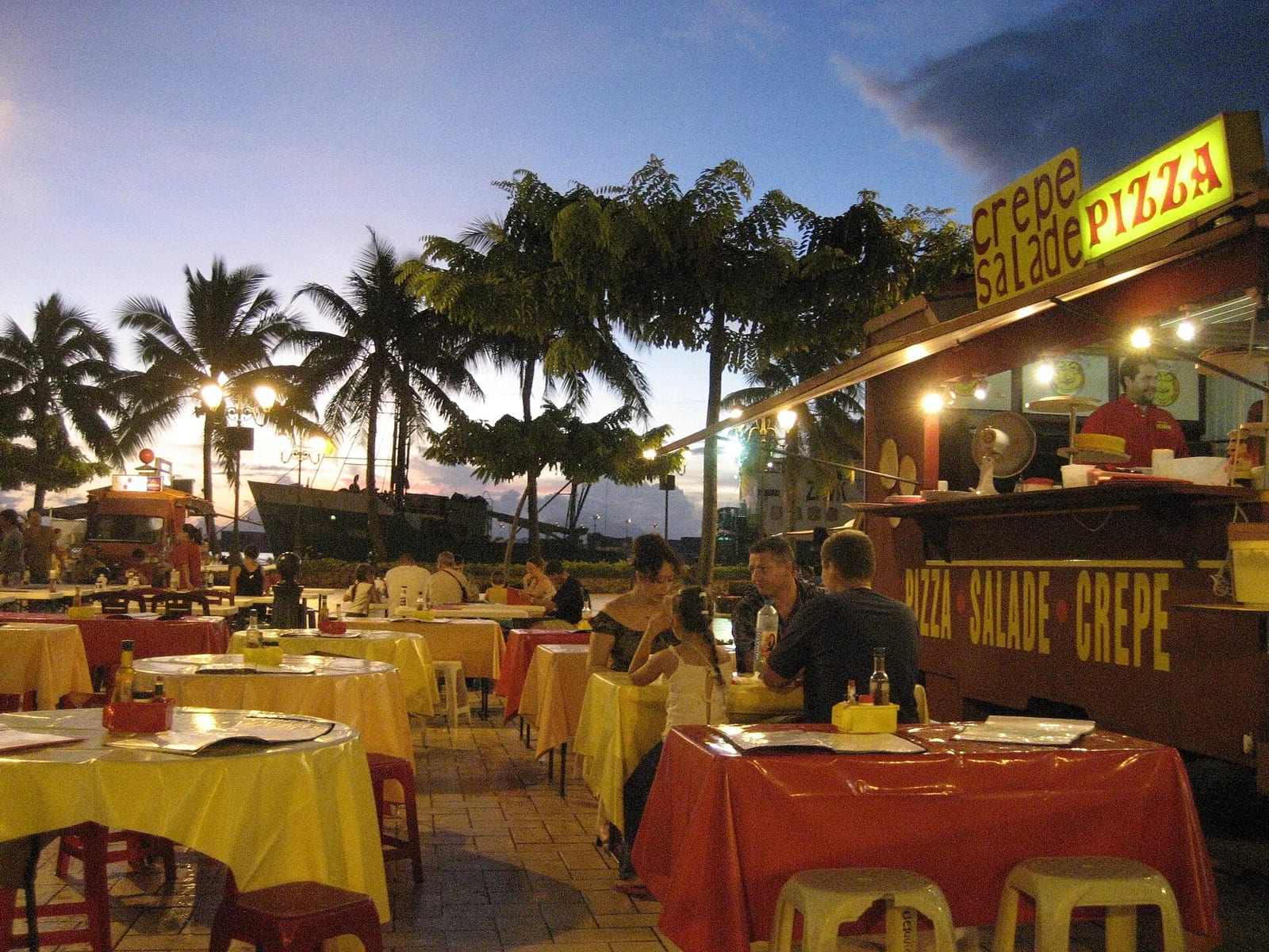 Roulottes au crépuscule en Polynésie : tables, tabourets et camion-snack crêpe salade pizza sous les palmiers
