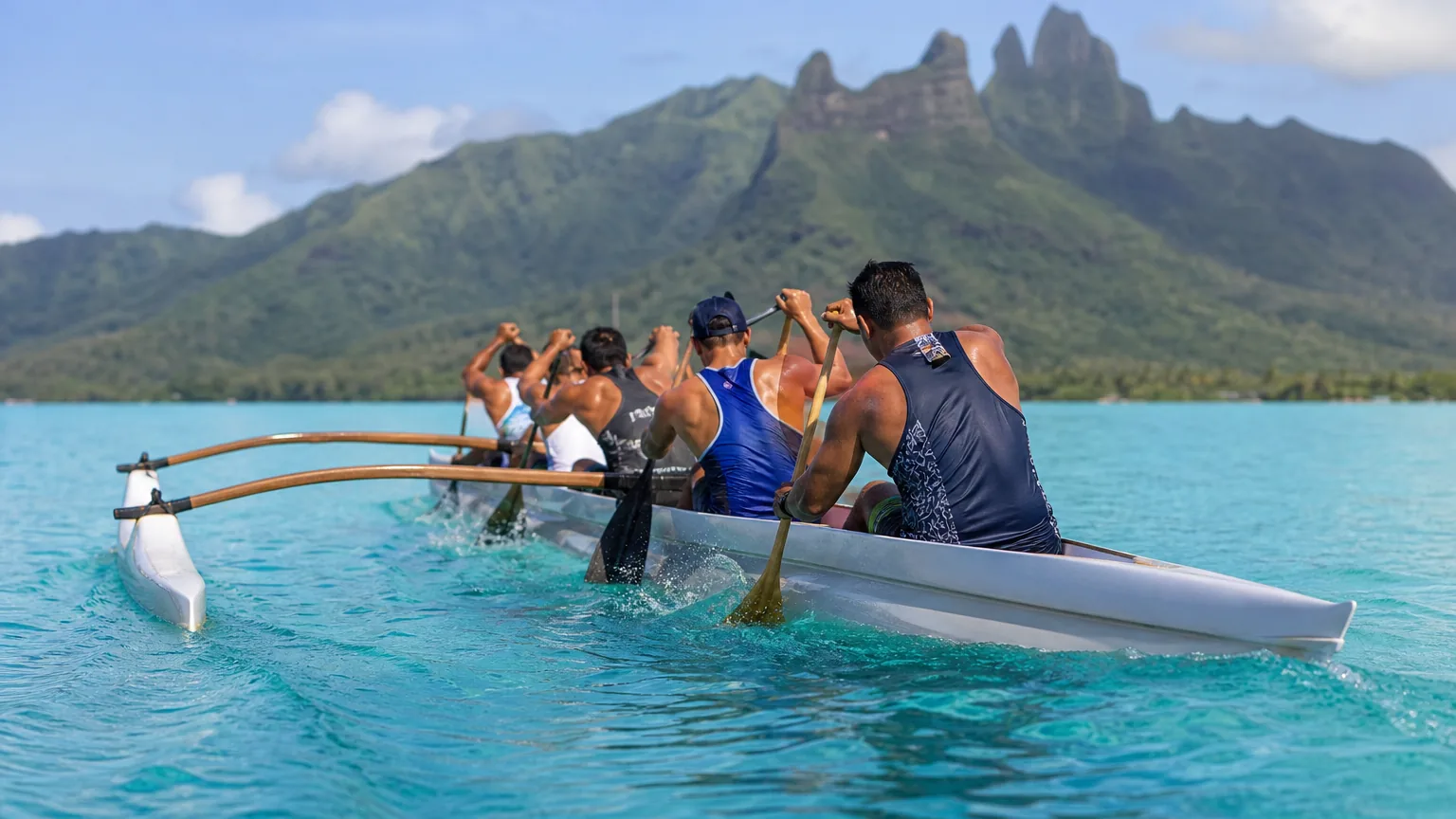 Équipage de va'a V6 pagayant en cadence sur un lagon turquoise, devant une montagne verte d'île haute