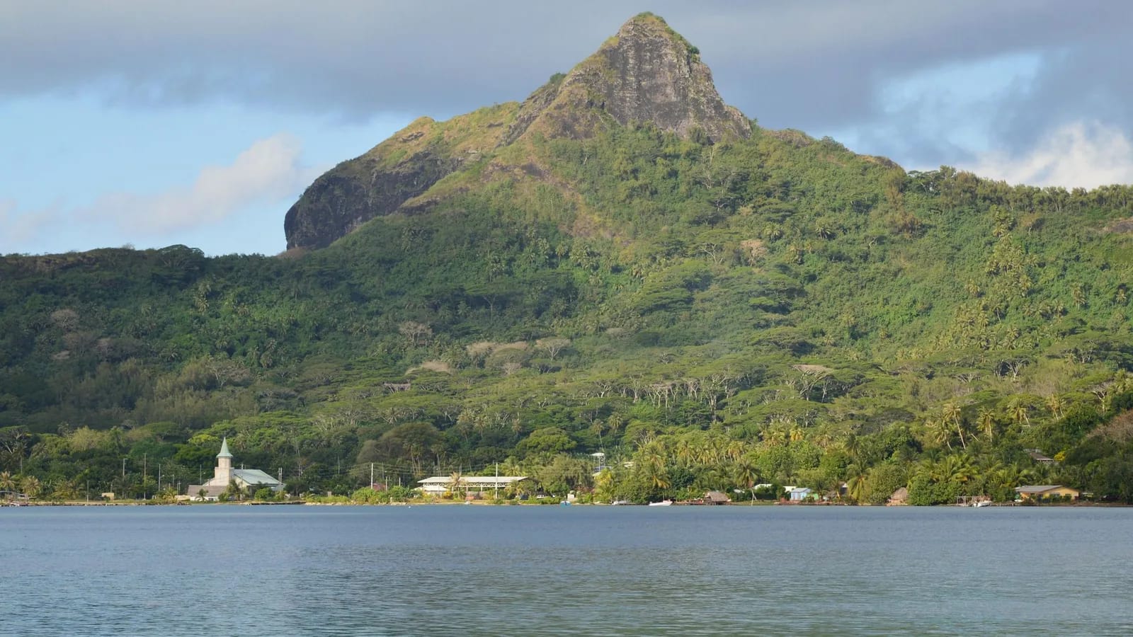 Montagne verdoyante et village avec son église au bord d'un lagon calme, dans les Îles Sous-le-Vent